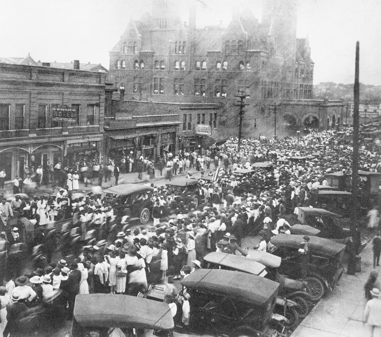 As a parade makes its way down Broadway, thousands of people gather in front of a hotel, shops and the city’s train station to watch. Parade participants are riding in and walking beside automobiles of the period.