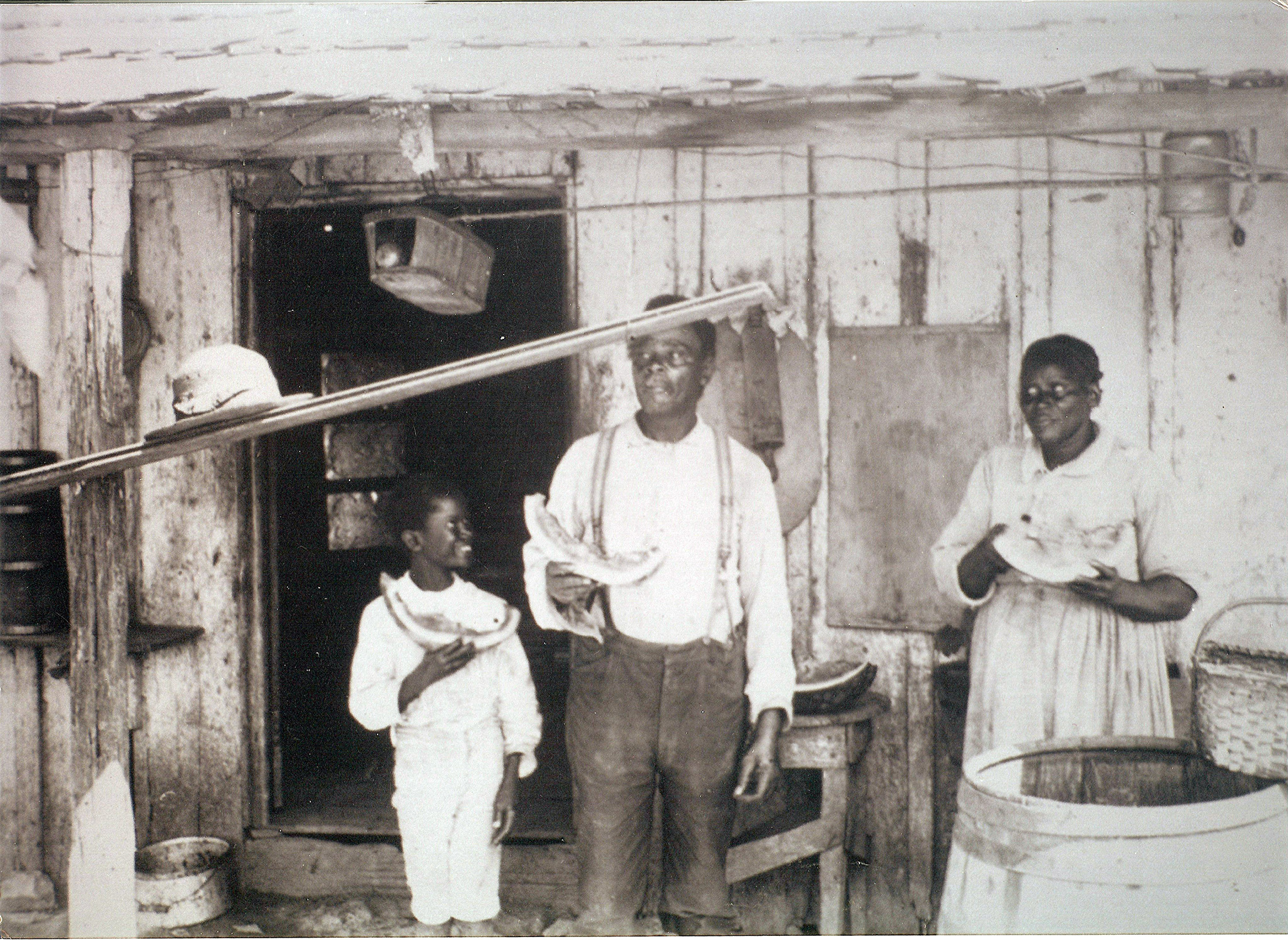 Black family standing outside their old wood house eating watermelon
