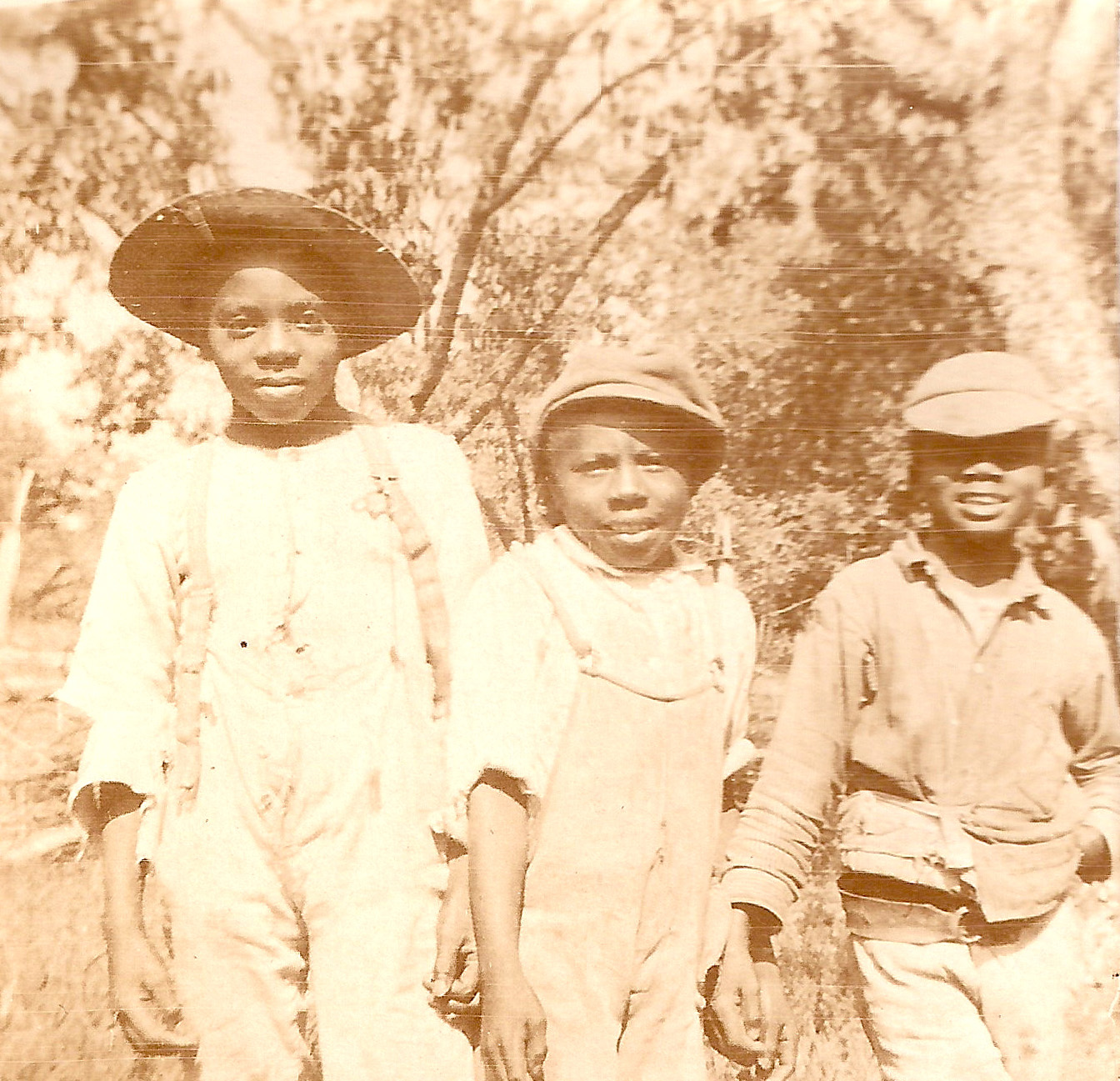 Sepia tone photograph of three young Black boys dressed in overalls and hats and smiling at the camera.