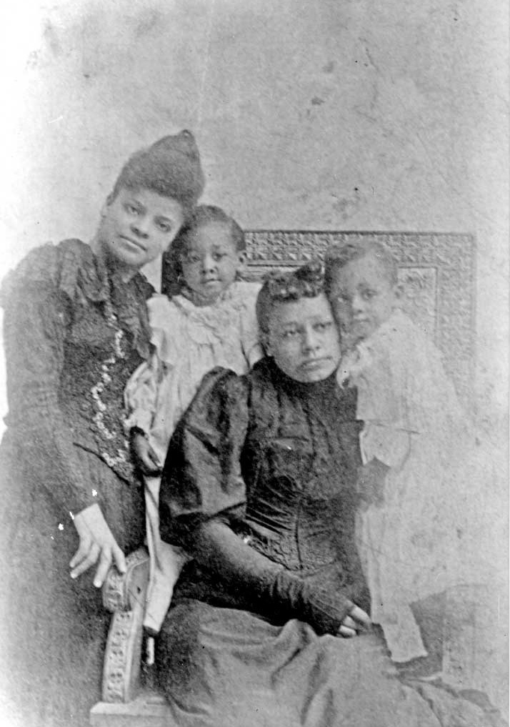 Old black and white photograph of two women dressed up and looking solemn with two younger children in dresses posing for the camera.