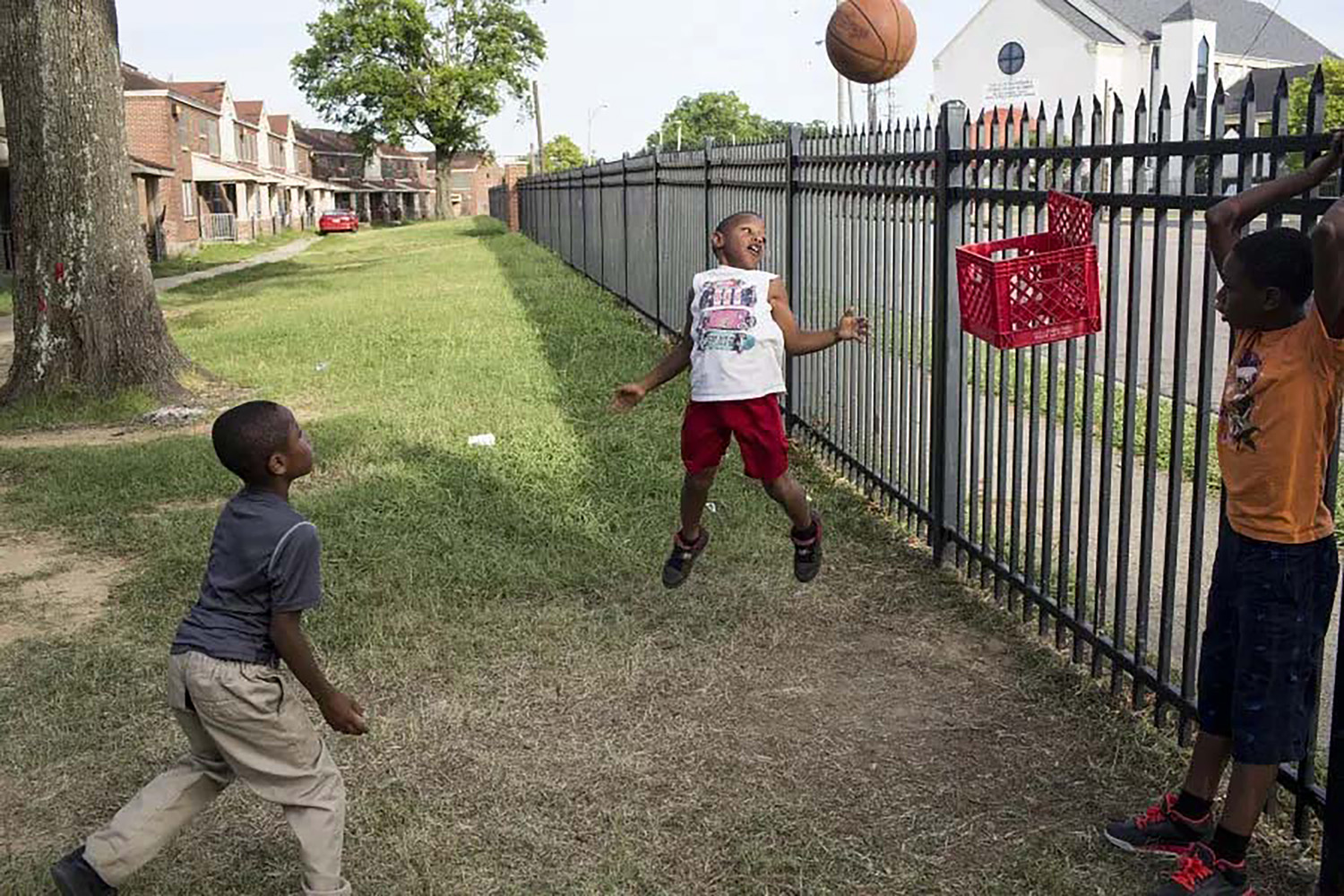 Three Black boys playing with a basketball on a grass lawn with a plastic bin attached to a fence acting as a basket.