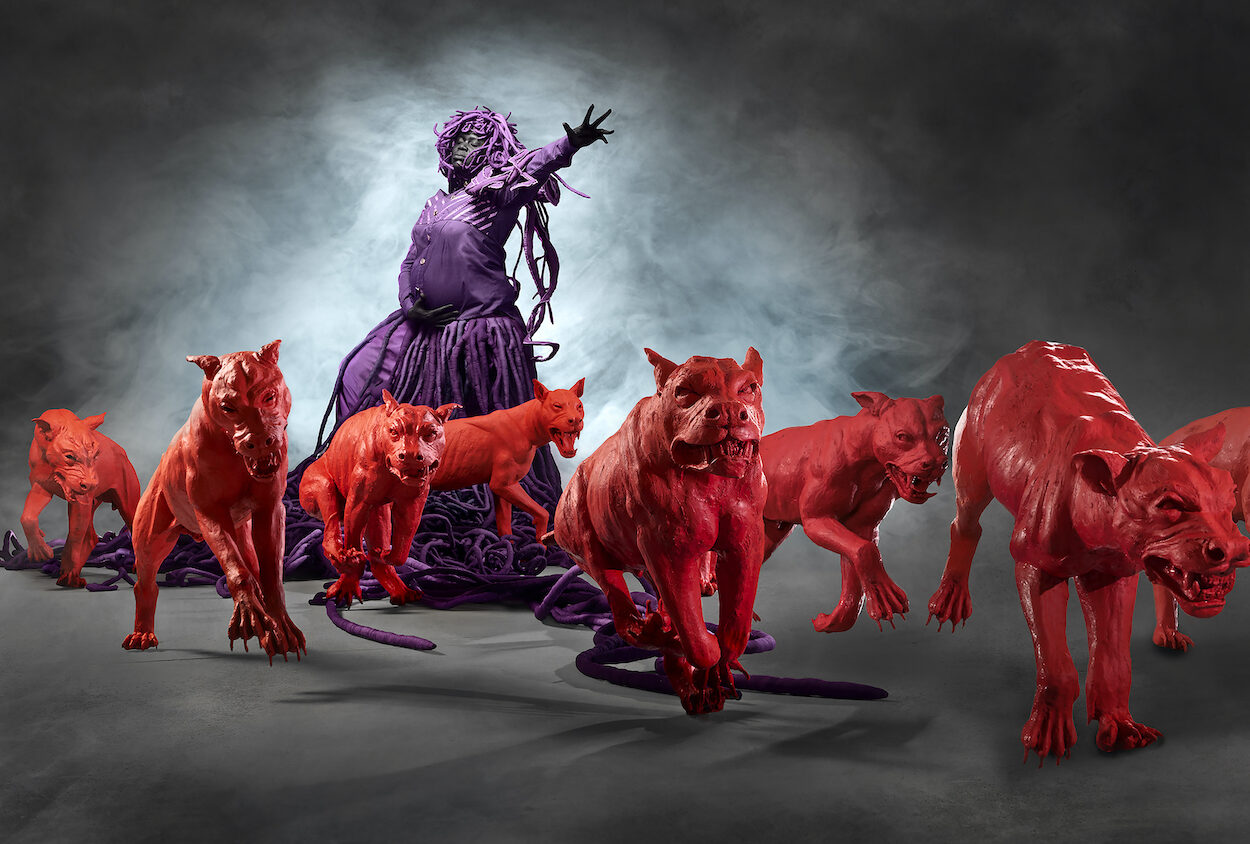 Wearing purple dress, a pregnant Black woman, eyes closed, cradles her belly with one arm. Her other arm outstretched with fingers splayed, gestures over a pack of vicious red dogs. Her long dreadlocks pool around her feet.