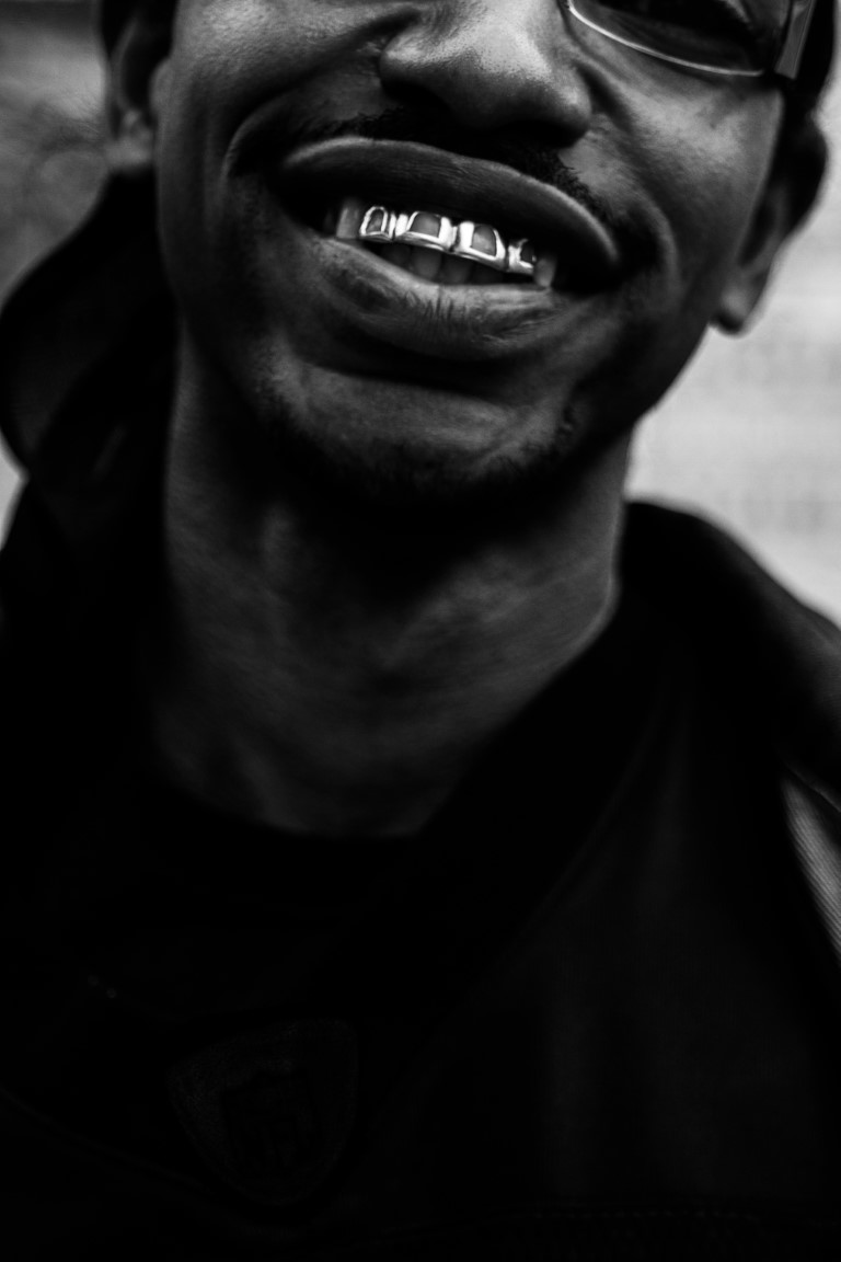 A black and white close-up of a black man in glasses smiling with a grill covering his top row of teeth.