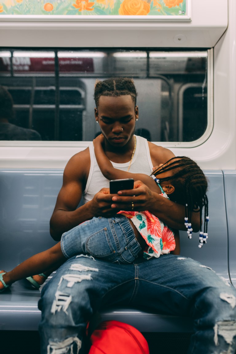 A black man riding a bus looks at his phone as he gently cradles a sleeping young girl in his lap. Her arm is tenderly around his neck.