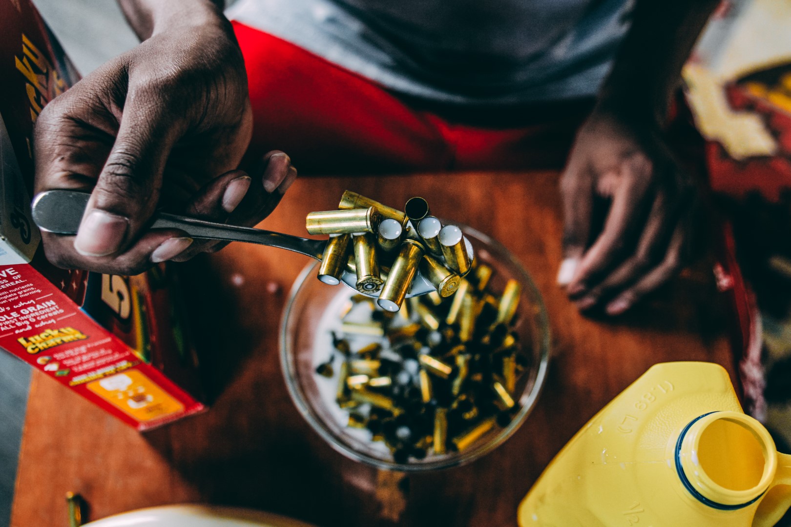 Overhead shot of a man in red shorts holding a spoon full of bullet shells from a glass bowl of milk. There’s a cereal box in the left corner and a yellow, gallon of milk in the lower, right corner.