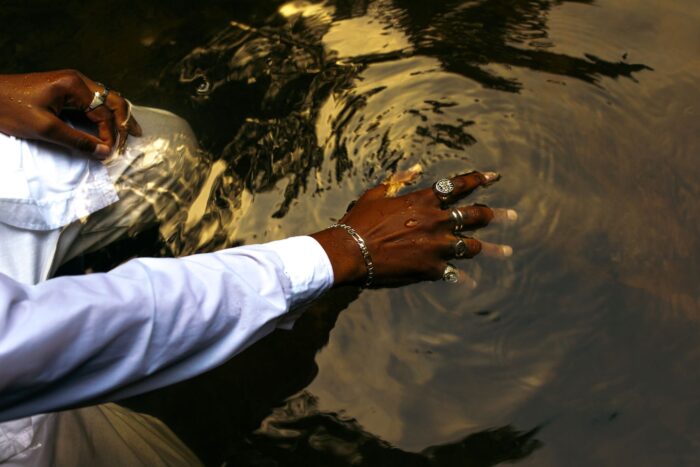Image of a man wearing a white, button-down dress shirt holding his hand out, kneeling in a shallow body of water. There’s a ring on each finger and a bracelet on his wrist.