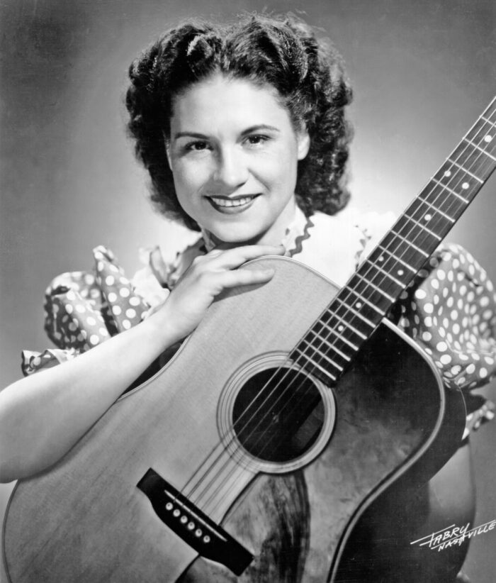 Black and white image of Kitty Wells smiling directly at the camera holding a guitar