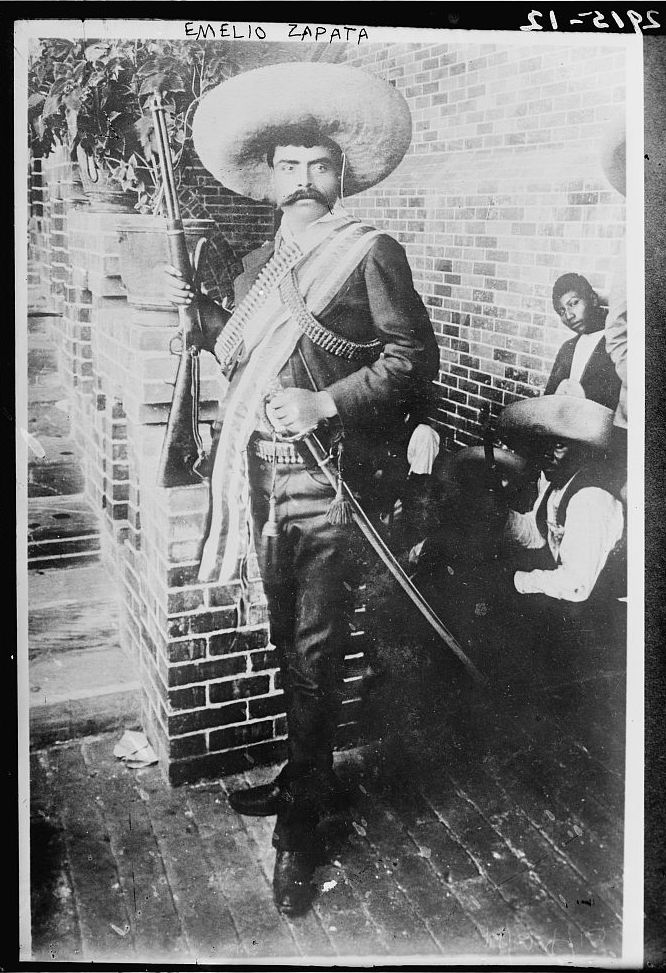 Emelio Zapata standing with a rifle in one hand and his hand on a sword in the other wearing a large brim hat