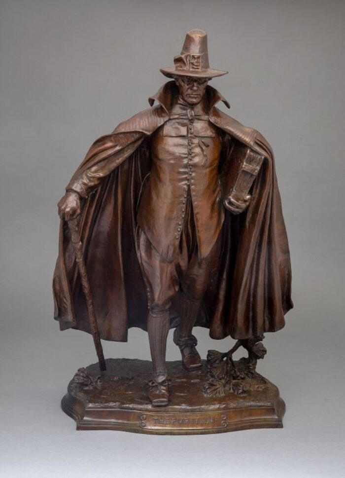Sculpture of a man wearing a long cape and tall hat walking sternly with a cane and a large book under his arm