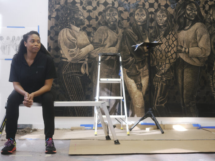 Artist LaToya Hobbs sitting on a chair next to some of her artwork.