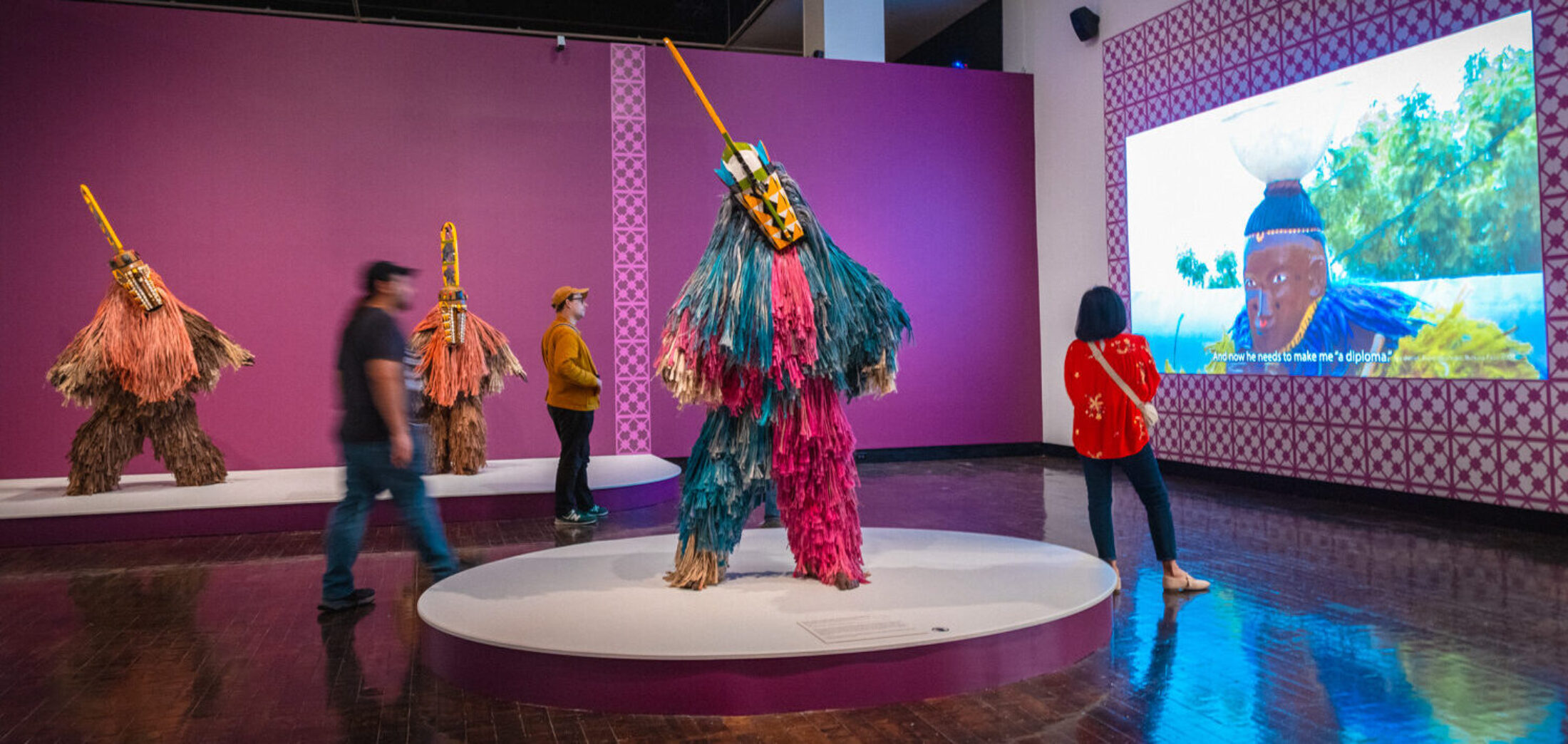 People in a gallery with large colorful masquerades and a a large video wall