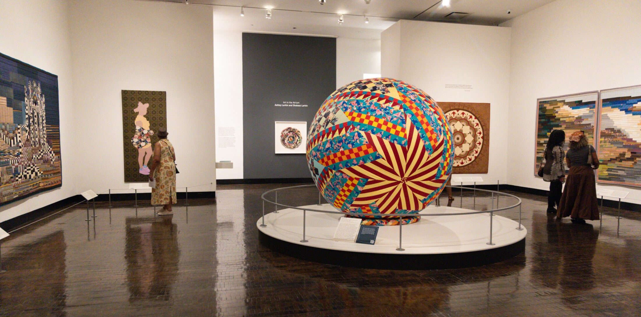 Wide view of people walking through a gallery with various quilts and a large quilt ball in the middle of the room