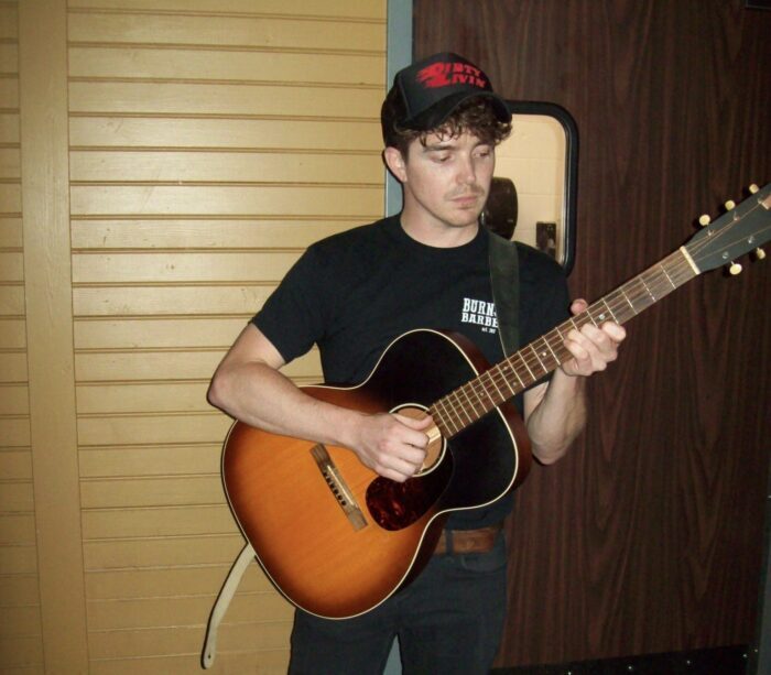 Musician Corey King stands with his guitar in front of a wooden door