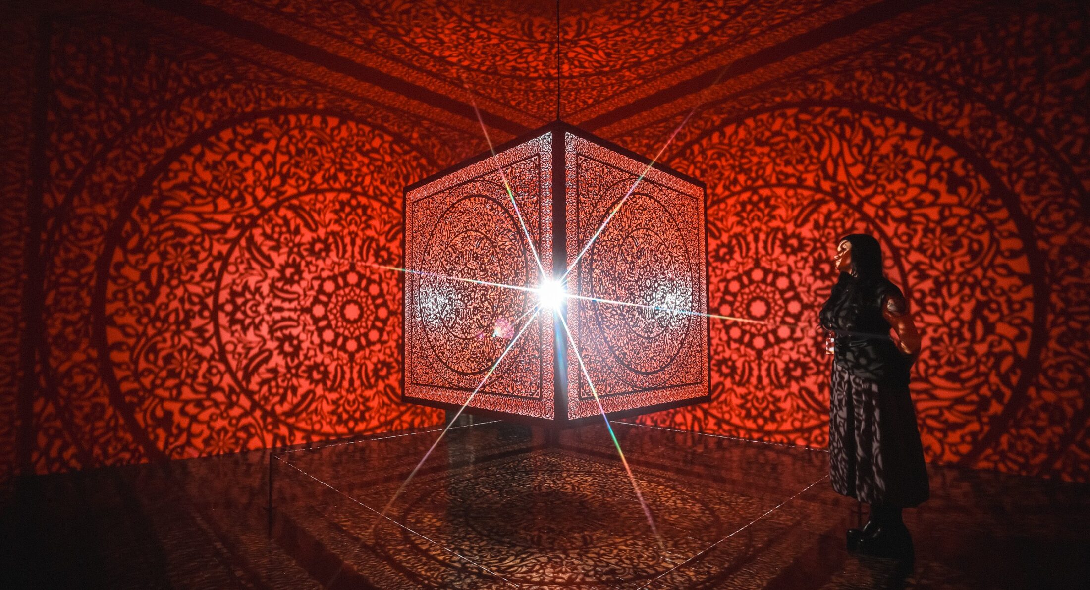 Woman standing in a room with a red cube handing in the center and red reflections on the walls.