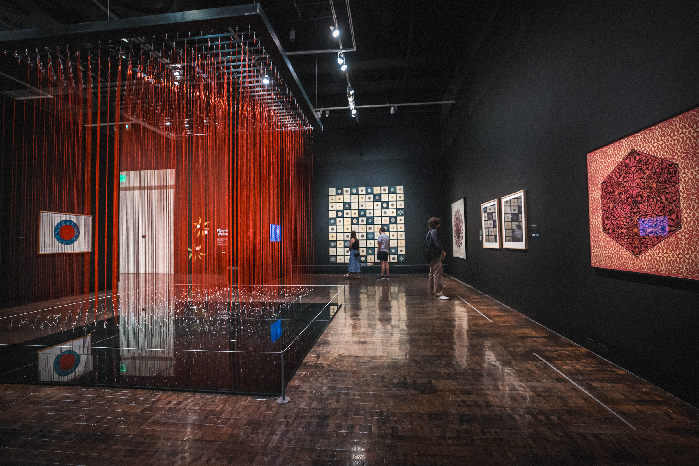 Gallery with an installation with tall red string-like pieces handing together in a square with other works on the walls