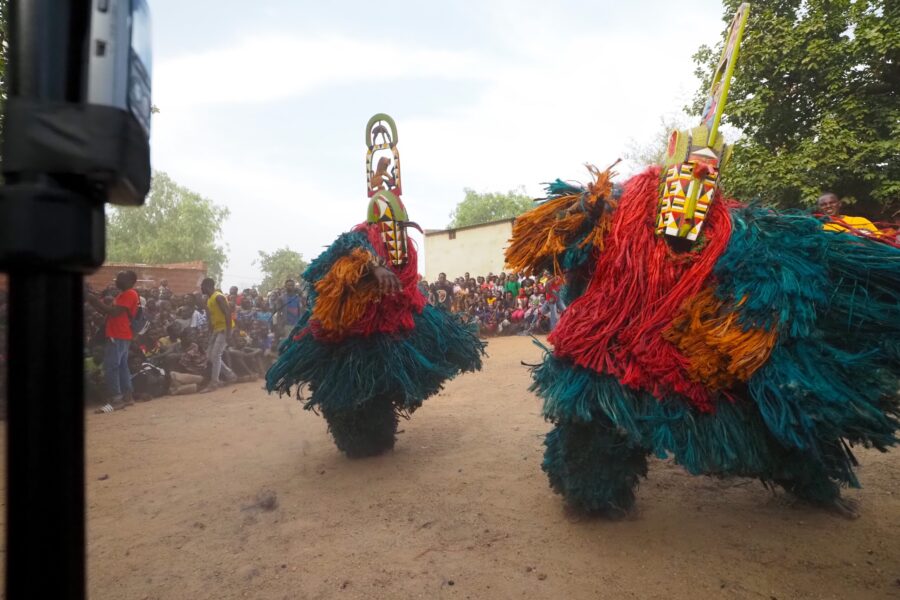 A pair of Kimi masks (headpiece carved by David Sanou in the studio of André Sanou) performing greetings with the lead griot Tchiedo playing his drum behind them.