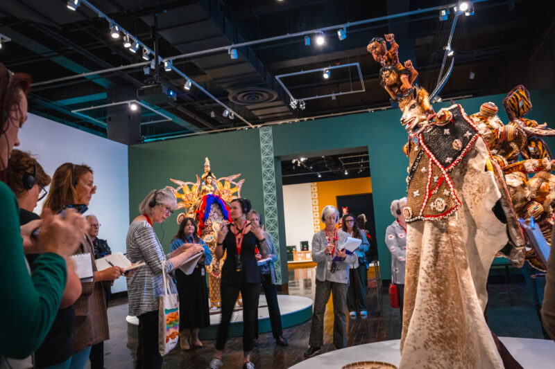 A woman leads a tour through a gallery with African Masquerades.