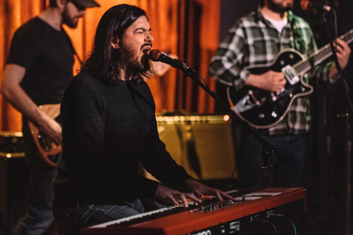 A man plays piano and sings with a band behind him.