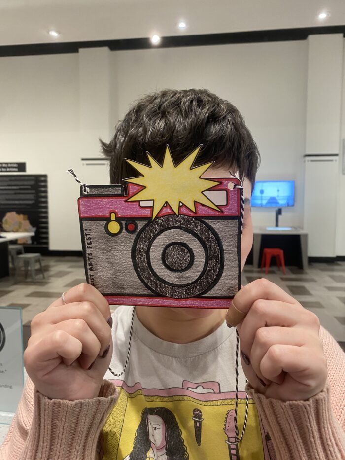 Woman holds a cut-out drawing of a camera like she's taking a photo.