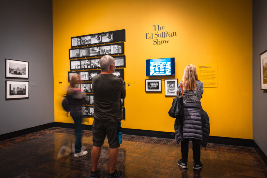 Three people look at photos in an art gallery with a bright yellow wall.