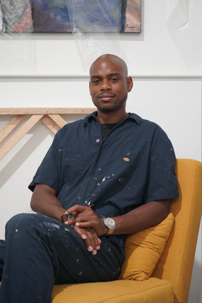 Artist Omari Booker poses for a photo on a yellow chair in an art studio.