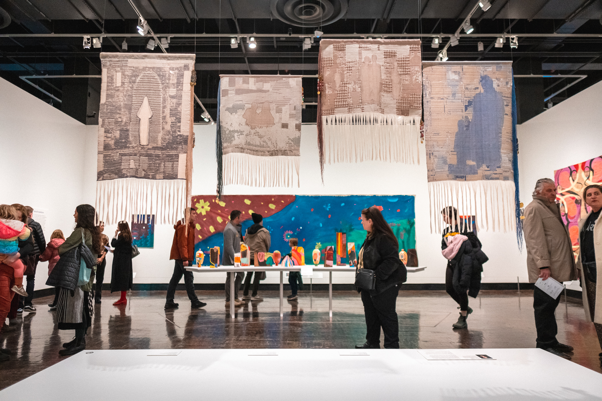 People walking through a large gallery with paintings on the walls and textiles hanging from the ceiling