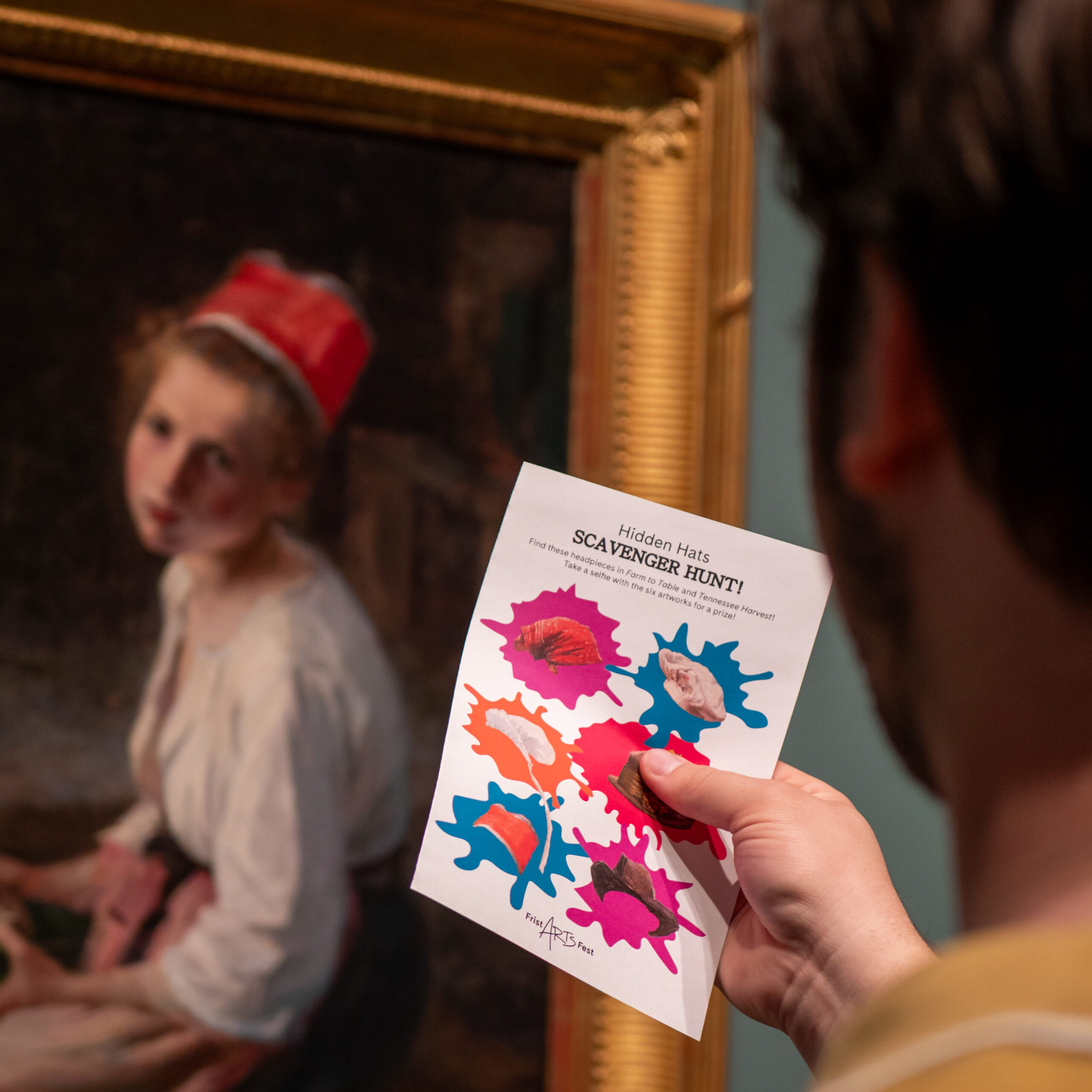 A man holds up a scavenger hunt card in front of a painting. 
