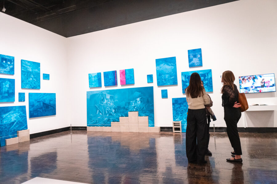 Three people have a conversation in front of blue paintings in an art gallery.