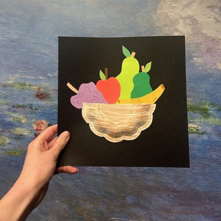 A hand holds a piece of black paper with fruit cut out of paper on it.