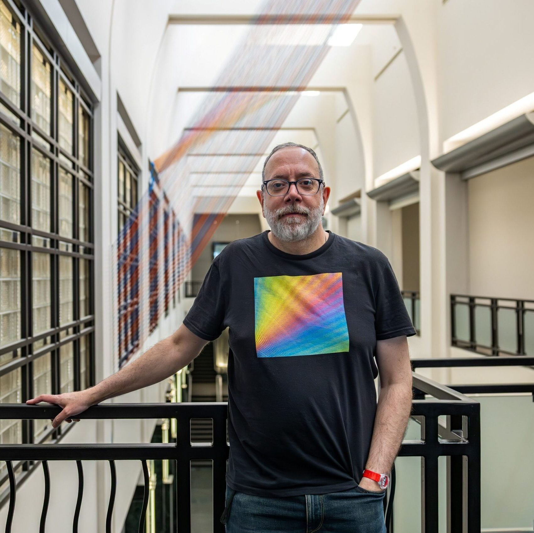 Gabriel Dawe poses in front of the Frist Art Museum upstairs balcony wearing a black shirt with a rainbow design on it.