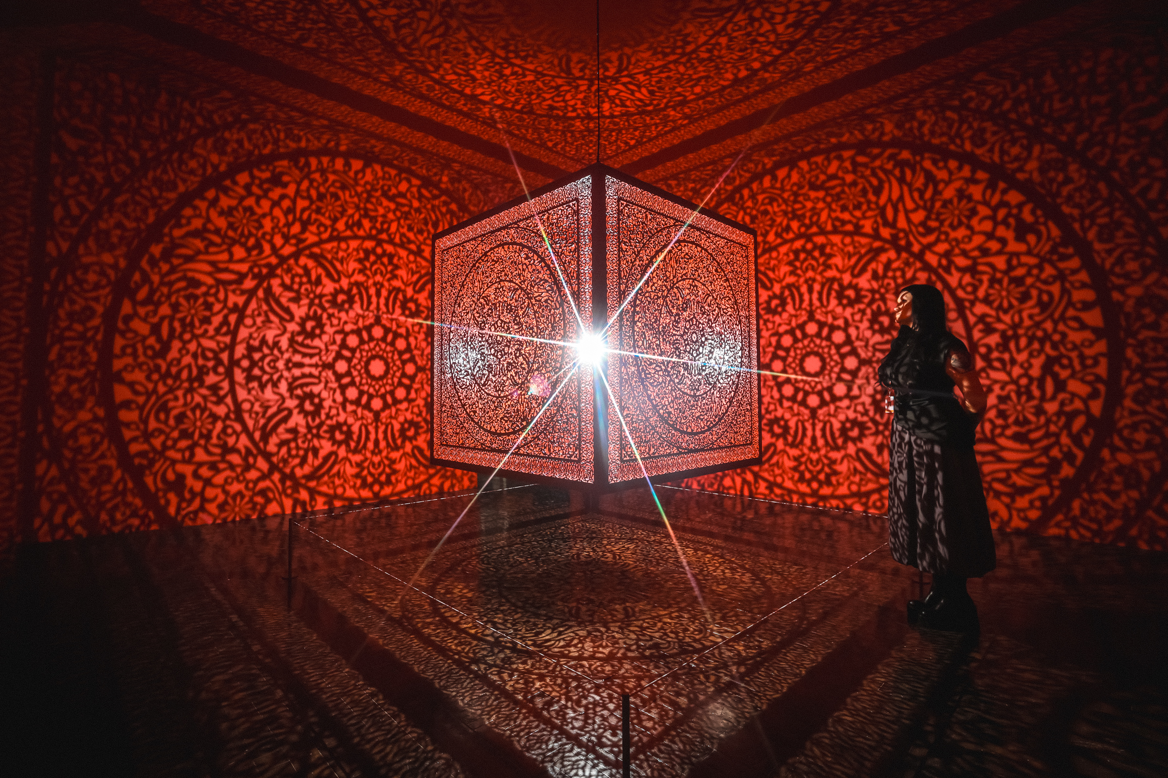 Woman standing in a room with a red cube handing in the center and red reflections on the walls.
