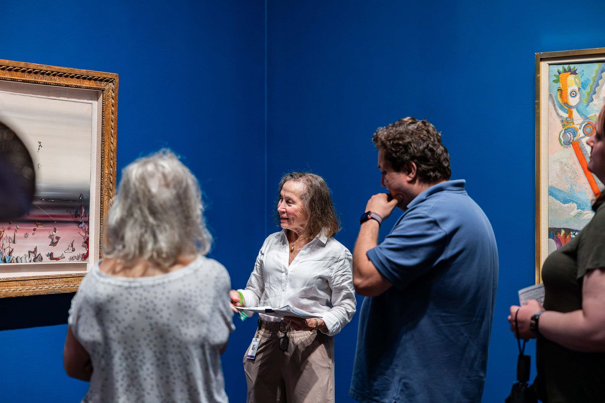 A woman gives a tour of an art gallery to a group of people in a room with blue walls.