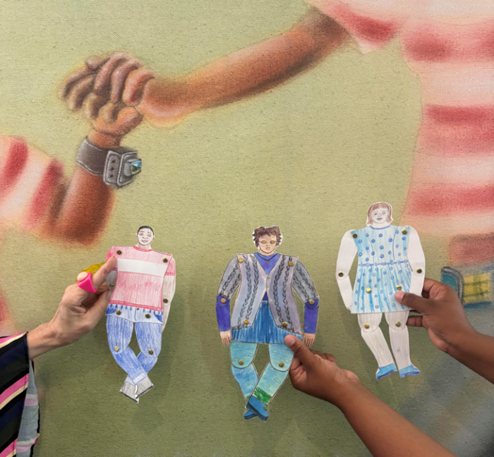 Three hands hold up paper puppets in front of an art piece with people holding hands. 