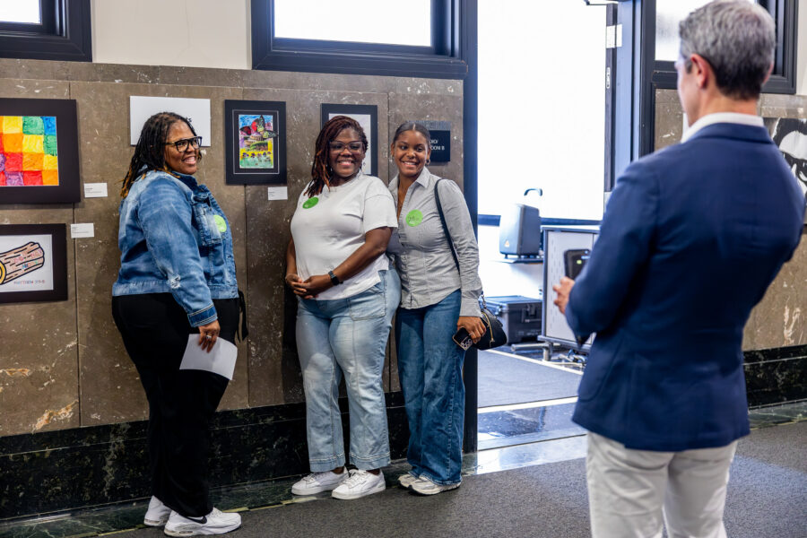 Three women pose for a photo in front of their art pieces in an art gallery.