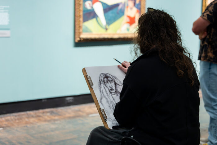 A woman with curly brown hair draws a picture of a painting. 