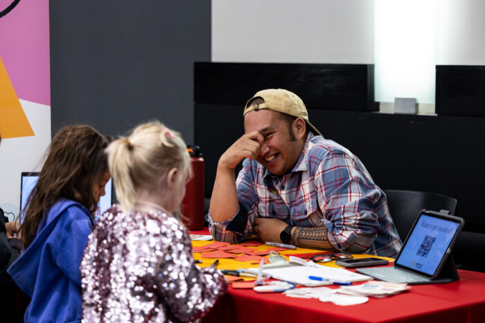 A man talks to two girls at a booth at a festival.