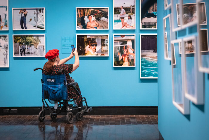 A woman in a wheelchair takes a photo on a phone of photographs in an art gallery.  