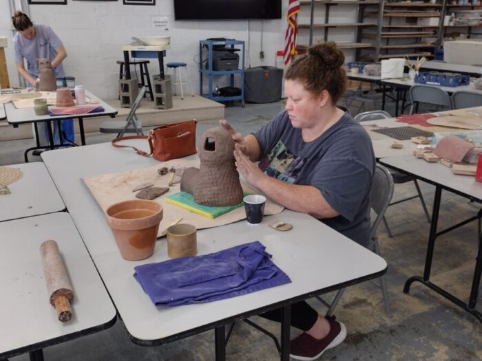 A woman sits at a table and creates a pottery sculpture.