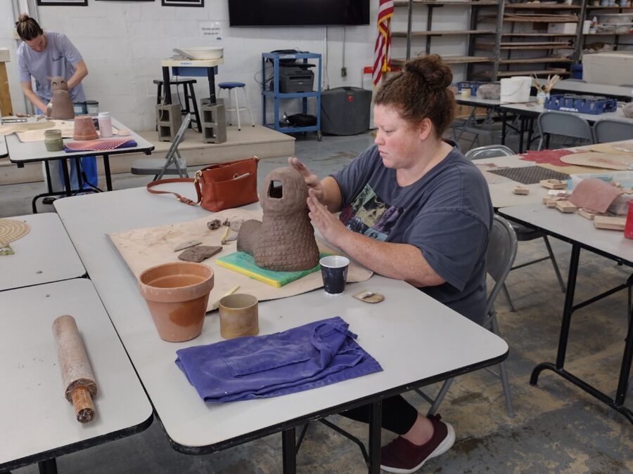 A woman sits at a table and creates a pottery sculpture.