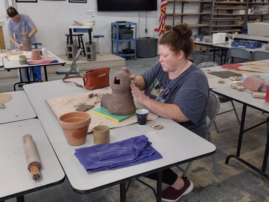 A woman sits at a table and creates a pottery sculpture.