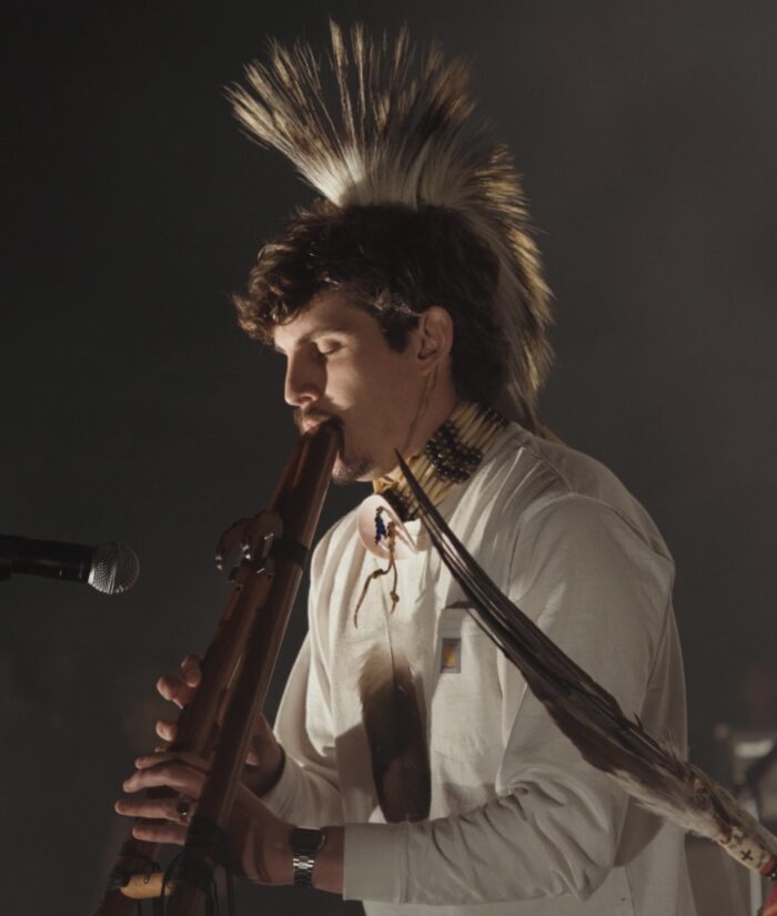 A man with a feather headpiece plays a woodland-style flute. 