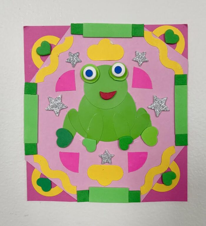 A crafted green frog with a patterned background in pink, green and yellow made out of paper.
