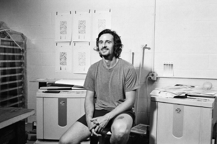 A black and white photo of a man sitting in a studio with art prints behind him.