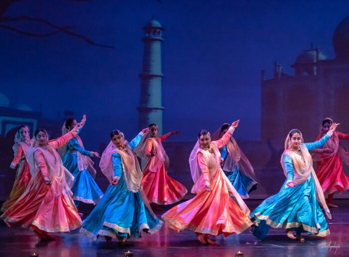A group of woman perform a dance dressed in traditional Indian clothing in colors of pink and blue. 