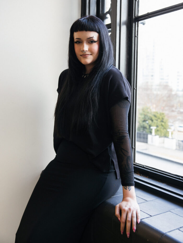A woman with black hair in an all black outfit leans on a window.