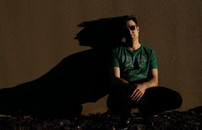 A heavily shadowed man in a green shirt and sunglasses leans against a wall.