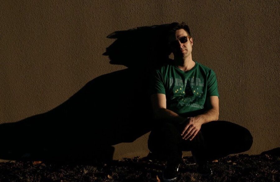 A heavily shadowed man in a green shirt and sunglasses leans against a wall.