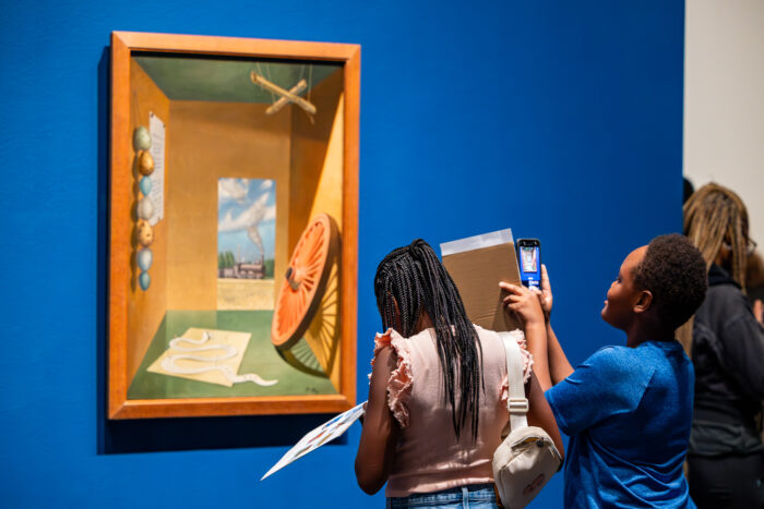 A young girl and boy take pictures of a painting in an art gallery with blue walls.