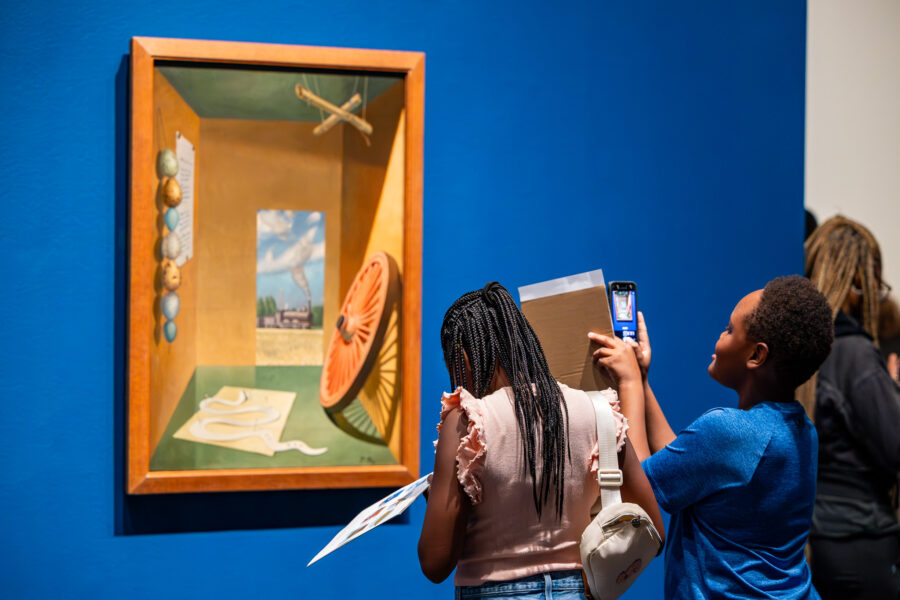 A young girl and boy take pictures of a painting in an art gallery with blue walls.