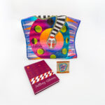 Bright colored tote bag with a snake on it next to a book and a patch on a white background.